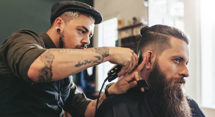 A man with a hat uses a black hair trimmer to trim the hair of a man with a long beard sitting in a chair.