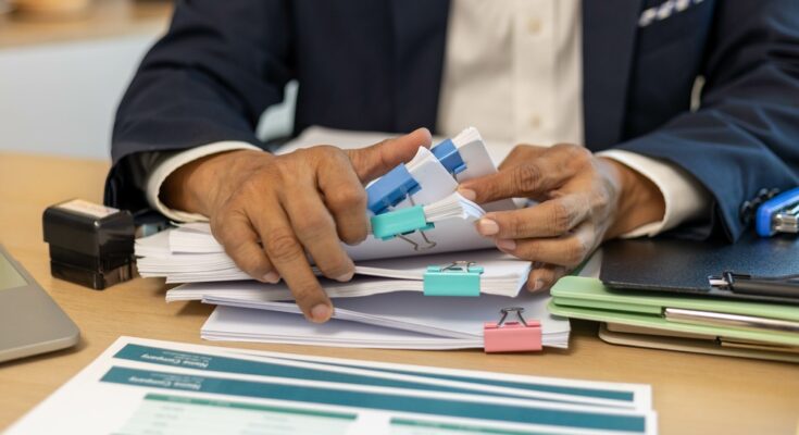 A man in business attire sorting through stacks of paper held together by binder clips of different colors.