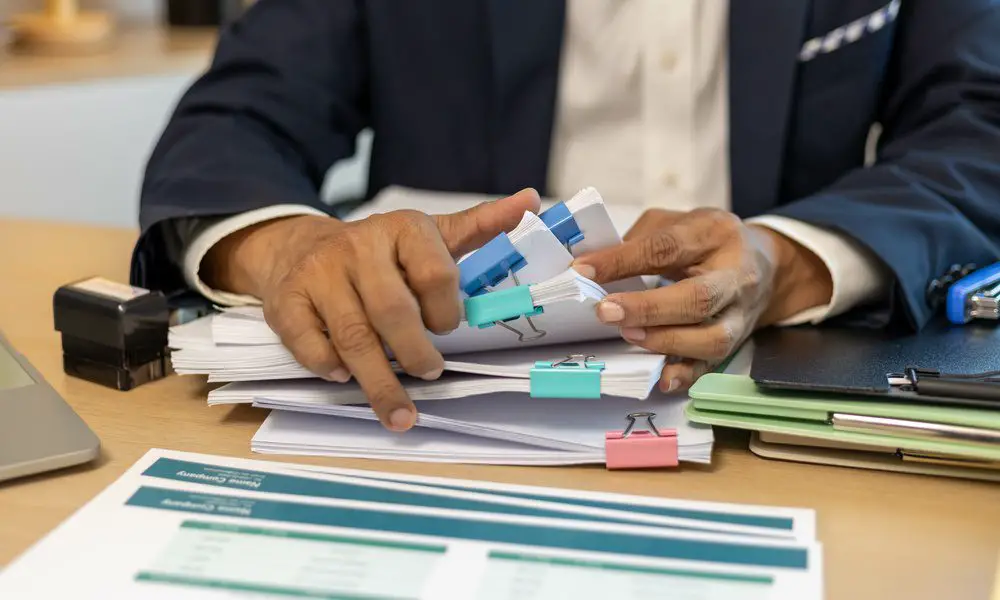 A man in business attire sorting through stacks of paper held together by binder clips of different colors.
