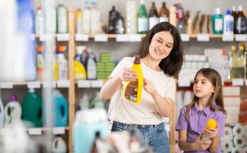A mother and young daughter stand in a store aisle filled with cleaning supplies. The mother points to a can of cleaner.